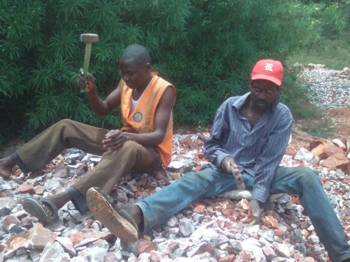 Charles Ouma, a Moi University student (left) in orange jacket and his dad (John Origi) in red cap, crushing concrete at a quarry site in Budalangi, Busia County, to raise money for fees. Despite the efforts, he has been unable to get the fees and appeals to people of goodwill to chip in and help him out. PHOTO/Gilbert Ochieng, The Scholar Media Africa.